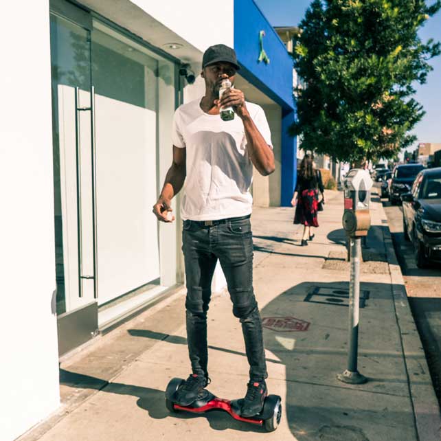 Man drinks juice on red hoverboard on sidewalk.